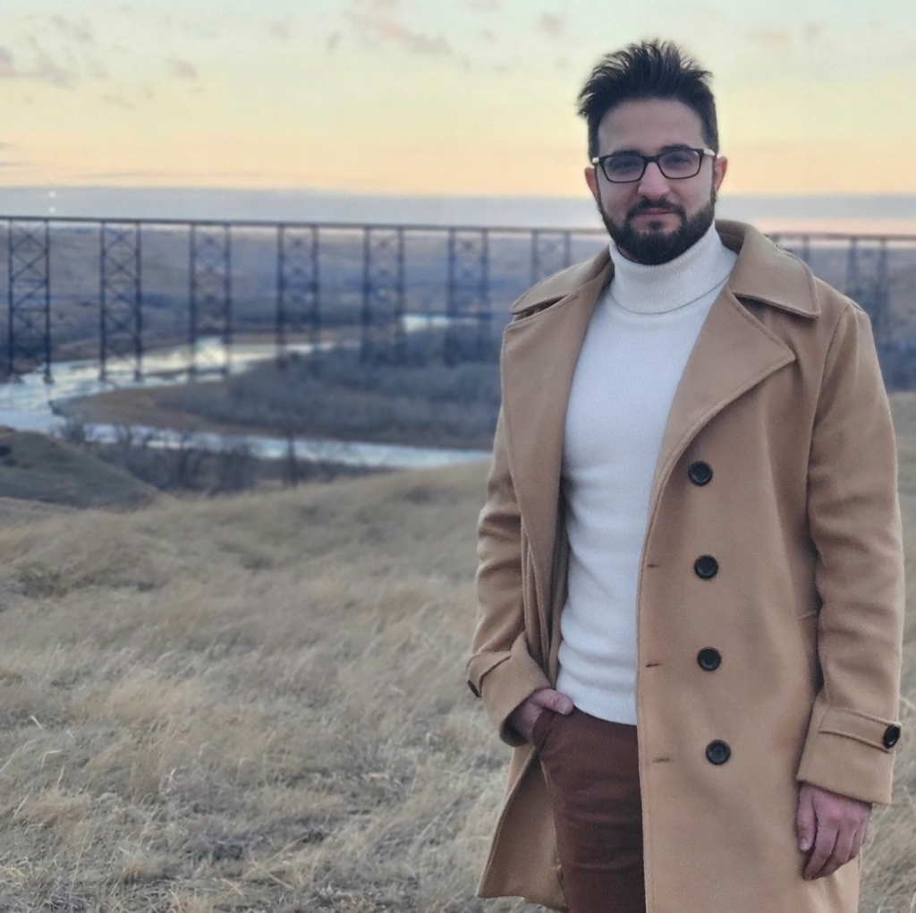 Dr. Amirhossein Moshrefi stands overlooking a large river valley with a bridge in the background. He is wearing a white sweater and tan coat, and has short dark hair and a trimmed beard.
